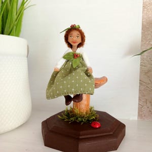 May include: A handmade doll with red hair and a green dress sits on a mushroom-shaped stand. The doll has a white top and brown shoes. A small red ladybug sits on the brown wooden base, which is decorated with moss.