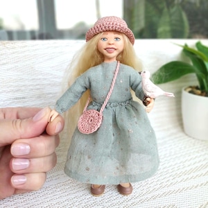 May include: A handmade doll with long blonde hair, wearing a light green dress, a pink crocheted hat, and a matching purse. The doll is holding a white bird. The doll's shoes are brown.
