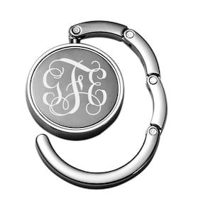 May include: A silver-toned metal purse hook with a round, engraved disc featuring a monogram of the letters "J F E" in a cursive script.