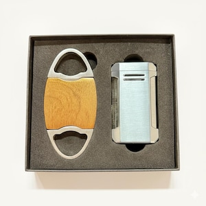 May include: A cigar cutter with a wooden center and silver-tone metal handle, and a silver and blue lighter, presented in a black box. The cutter has a rounded, oval shape. The lighter has a rectangular shape.