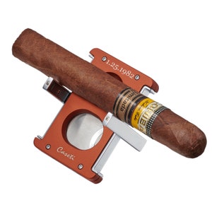 May include: A red and silver cigar cutter with a cigar resting in the cutter. The cigar is labeled "Romeo y Julieta" and "1.25.1982". The cutter is labeled "Caseti".