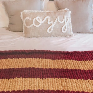 May include: A bed with white bedding and two throw pillows with a third pillow that says "cozy" in white lettering. A striped throw blanket with burgundy and tan stripes is on the bed.