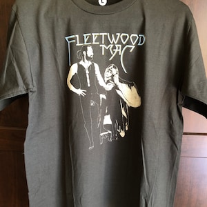 May include: A dark grey t-shirt with a white graphic of Fleetwood Mac. The graphic features two figures, a man and a woman, in a silhouette style.