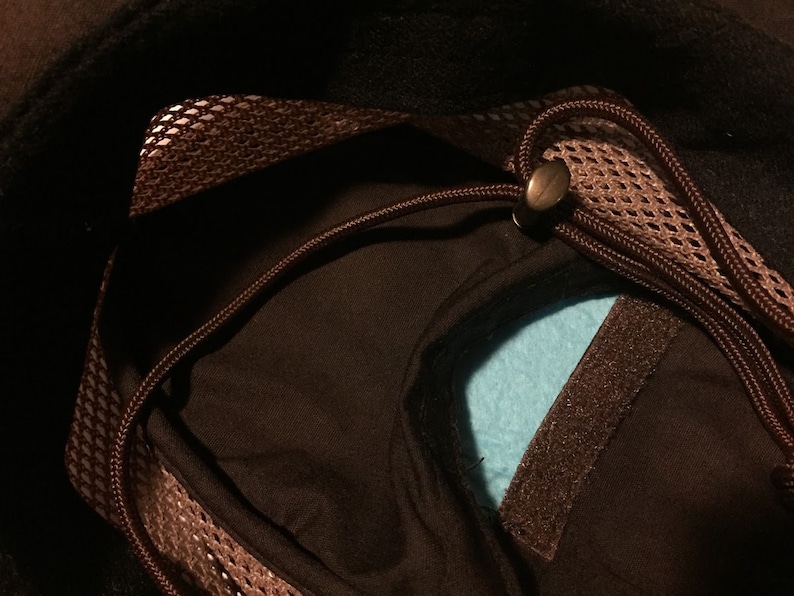 May include: Close-up of a black hat interior with a brown mesh trim and drawstring. A light blue fabric is visible inside the hat. A brown velcro strip is also visible.