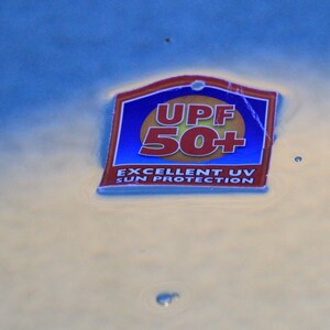 May include: A rectangular tag with a blue background and a red and gold border floats on water. The tag reads "UPF 50+" in red and "EXCELLENT UV SUN PROTECTION" in white.