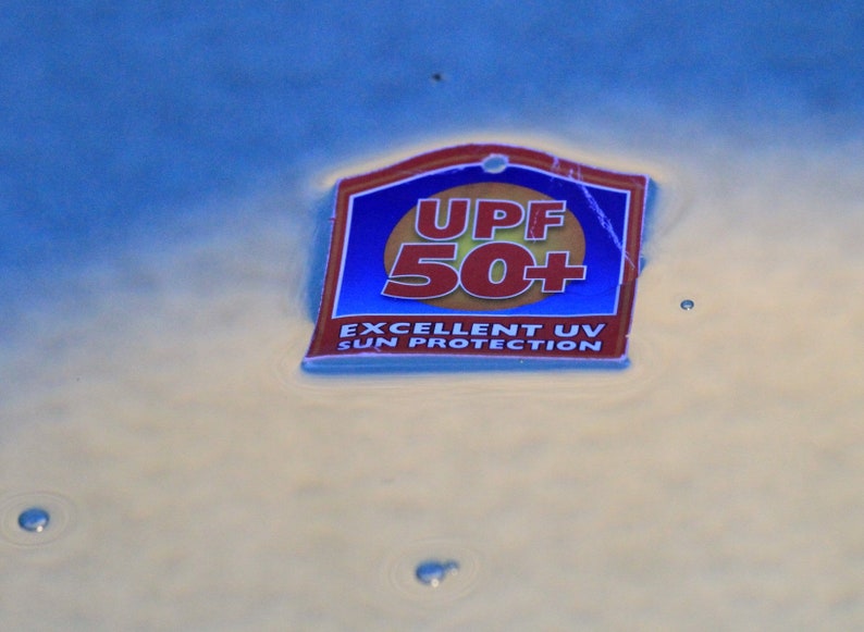 May include: A rectangular tag with a blue background and a red and gold border floats on water. The tag reads "UPF 50+" in red and "EXCELLENT UV SUN PROTECTION" in white.