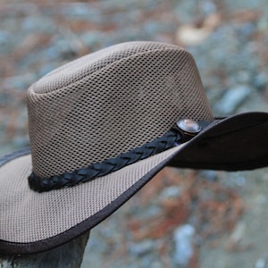 May include: A brown mesh cowboy hat with a dark brown brim and a decorative band. The hat features a metal button and a braided leather band around the crown. The hat is designed for sun protection.