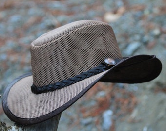 Könnte beinhalten: Brauner Cowboyhut aus Mesh mit dunklem braunem Rand und Zierband. Der Hut hat einen Metallknopf und ein geflochtenes Lederband um die Krone. Der Hut ist für den Sonnenschutz konzipiert.