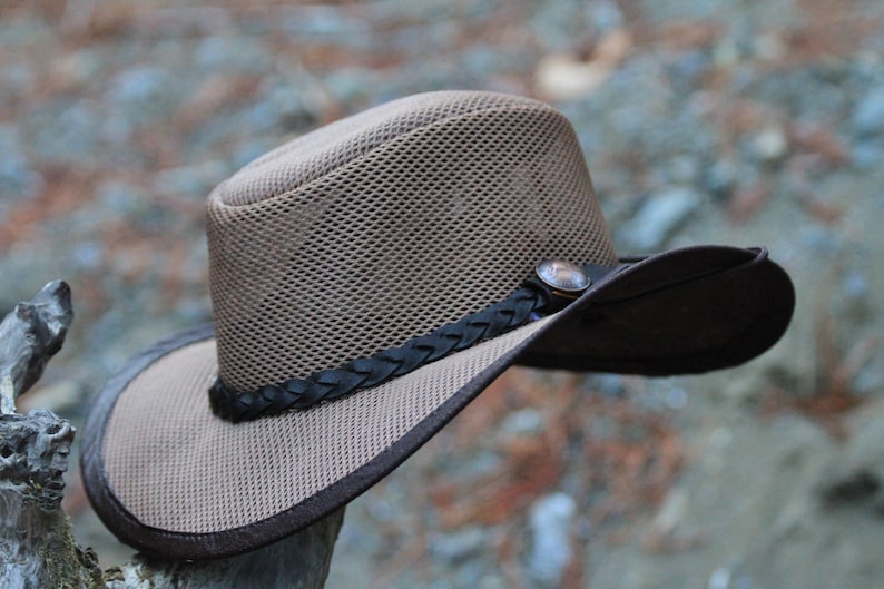 May include: A brown mesh cowboy hat with a dark brown brim and a decorative band. The hat features a metal button and a braided leather band around the crown. The hat is designed for sun protection.