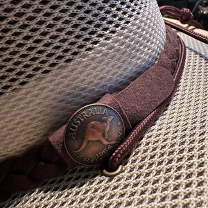 May include: Close-up of a mesh-textured hat with a brown leather band and a copper-colored coin. The coin features a kangaroo and the word "Australia" with the year 1964. The hat has a braided cord and a metal ring.