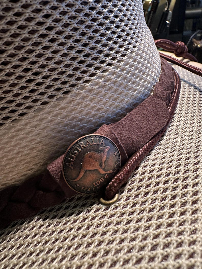 May include: Close-up of a mesh-textured hat with a brown leather band and a copper-colored coin. The coin features a kangaroo and the word "Australia" with the year 1964. The hat has a braided cord and a metal ring.