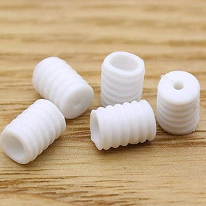 May include: Five small, white, cylindrical plastic beads with a ridged design. The beads are scattered on a light brown wooden surface. One bead has a hole through the center.