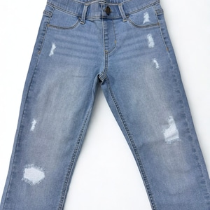 May include: Light blue denim jeans with distressed details. The jeans have a classic five-pocket design, a button closure, and a faded wash. The jeans are laid flat on a white surface.