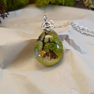 May include: A teardrop-shaped pendant necklace with a silver chain. The pendant is filled with clear resin and contains a small green tree, gold flakes, and iridescent glitter.