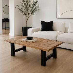 May include: A rectangular coffee table with a natural wood top and black legs. A black bowl and a book are on the table. The table is in a living room with a white sofa and a black pillow.