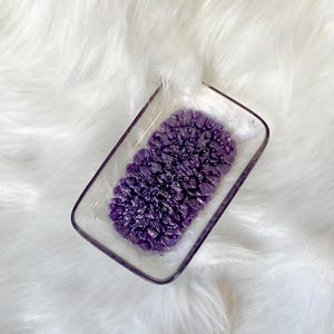 May include: A small, rectangular dish made of clear glass with a purple, textured design inside. The dish is sitting on a white, fluffy surface.