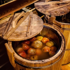 May include: Wooden barrels with lids, one partially open, revealing tomatoes in a liquid. The barrels are made of light brown wood and have metal bands. The lids are round with a handle.