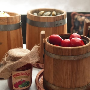 May include: A collection of wooden barrels with metal bands, some filled with pickled vegetables and red tomatoes. A glass jar with a burlap-covered lid features a label with the text "ТРИПІЛЬСЬКІ РІДНОСОЛ" and illustrations of tomatoes and cucumbers.
