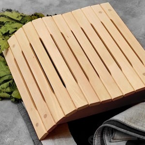 Può includere: Un poggiatesta per sauna in legno chiaro caratterizzato da un design curvo a doghe con assi parallele. Il poggiatesta è posizionato accanto a un mucchio di foglie di quercia verdi essiccate e un asciugamano in tessuto grigio con stampa di zampa.