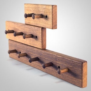 May include: A wooden wall-mounted coat rack with multiple hooks. The rack is made of three horizontal pieces of wood, each with a different number of hooks. The hooks are made of wood and are stained a dark brown colour.