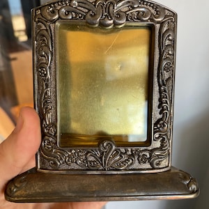 May include: A small, rectangular, antique silver-toned picture frame with ornate floral and scroll detailing. The frame has a built-in stand and a gold-colored interior. The frame is held by a hand.
