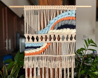 Pode incluir: Uma tapeçaria de macramé bege com um design tecido com fio azul, branco, laranja e cinza. A tapeçaria tem uma borda de franjas e está montada em uma vareta de madeira.
