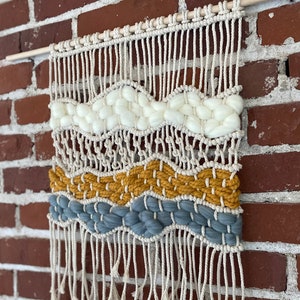 Puede incluir: Un tapiz de macramé de pared con un patrón blanco, amarillo y azul. El tapiz tiene una franja en la parte inferior y está hecho con fibras naturales.