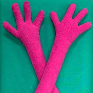 Puede incluir: Un par de calentadores de brazo de felpa rosa con extremos en forma de mano. Los calentadores de brazo están cruzados uno sobre el otro.