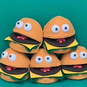 Puede incluir: Cinco hamburguesas de peluche con ojos saltones y bocas abiertas. Las hamburguesas están apiladas en forma de pirámide, con la hamburguesa superior mirando hacia adelante y las demás hacia afuera.