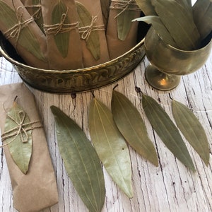 May include: A collection of dried bay leaves wrapped in brown paper and tied with twine, displayed in a gold metal bowl. Several loose bay leaves are scattered on a white wooden surface.