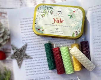 May include: A set of six small, colorful beeswax candles in green, red, light green, yellow, white, and purple. The candles are next to a decorative tin with the text "Yule" and "Tiny Candles for Mindful Rituals". The tin is on a book.