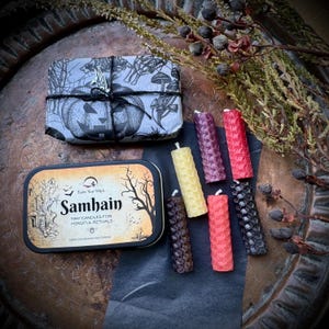 Peut inclure: Un ensemble de petites bougies colorées, dont jaunes, rouges et violettes, disposées avec une boîte décorative et un paquet emballé. La boîte porte l'inscription "Samhain" et "Tiny Candles for Mindful Rituals".