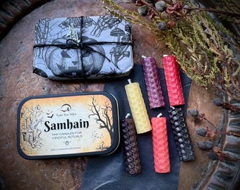 May include: A collection of tiny candles in various colors, including yellow, red, and purple, arranged with a decorative tin and a wrapped package. The tin reads "Samhain" and "Tiny Candles for Mindful Rituals."