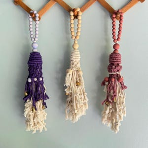 May include: Three decorative macrame tassels in purple, cream, and mauve, each with a beaded top and small gold bells. The tassels hang from a wooden rack against a light blue wall, showcasing a bohemian style.