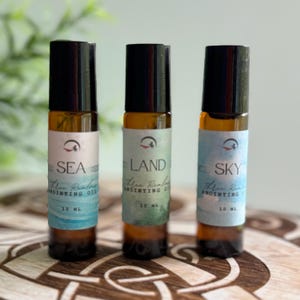May include: Three 10 ml amber glass roller bottles with black caps. Each bottle has a white label with a blue and green watercolor design. The labels read "SEA", "LAND", and "SKY" with the text "Three Realms Anointing Oil" underneath.