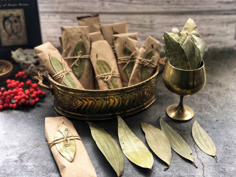 May include: A brass bowl filled with bundles of dried bay leaves wrapped in brown paper. The leaves are tied with twine and arranged in a decorative pattern. Several loose bay leaves are scattered on a gray surface.