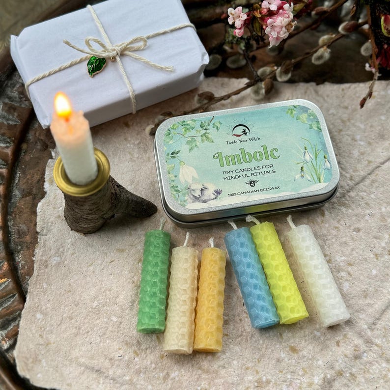 Może przedstawiać: Zapalone biała świeca w uchwycie z kory brzozy, białe pudełko prezentowe związane sznurkiem i puszka z napisem "Imbolc Tiny Candles for Mindful Rituals" są wyświetlane. Sześć małych, kolorowych świec z wosku pszczelego jest ułożonych z przodu.