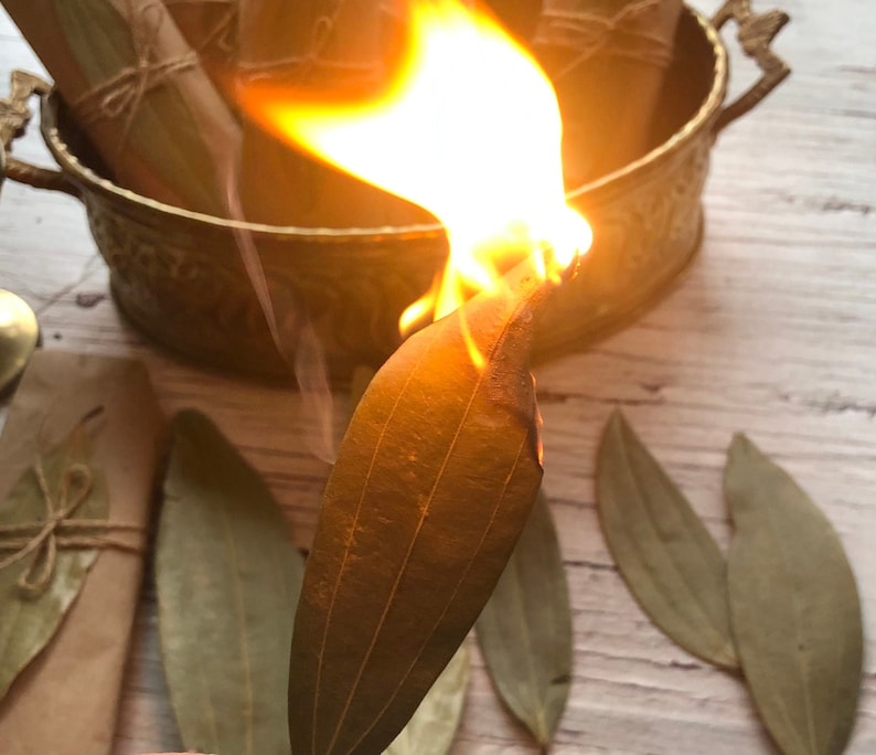 May include: A close-up of a bay leaf burning with flames. The leaf is held over a brass bowl. There are several other bay leaves on a white wooden surface.