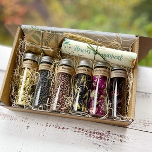 May include: A gift box filled with six glass test tubes containing dried flowers. The test tubes are labelled with the names of the flowers: Jasmine, Lavender, Heather, Butterfly Pea, Globe Amaranth, and Hibiscus. The box is decorated with a scroll that reads "Flowers are magical".
