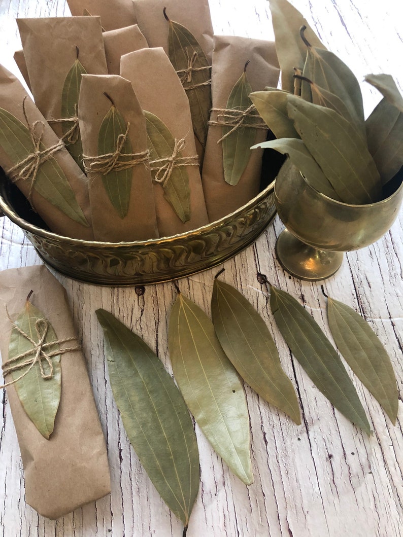 May include: A collection of dried bay leaves wrapped in brown paper and tied with twine, displayed in a gold metal bowl. Several loose bay leaves are scattered on a white wooden surface.