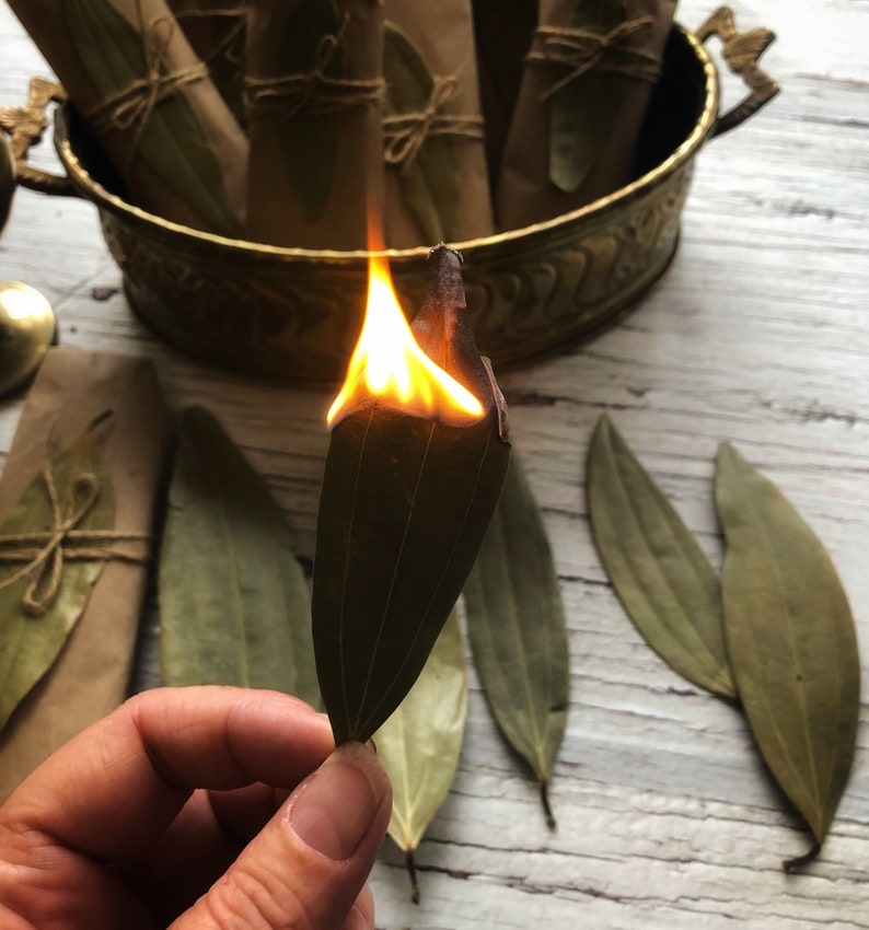 May include: A hand holds a dried bay leaf that is on fire. The leaf is surrounded by other dried bay leaves on a white wooden surface. A bowl of dried bay leaves is in the background.