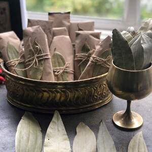 May include: A brass bowl filled with small brown paper packages tied with twine and decorated with dried bay leaves.  A gold goblet sits next to the bowl, also filled with dried bay leaves.  Several dried bay leaves are scattered on a gray surface.