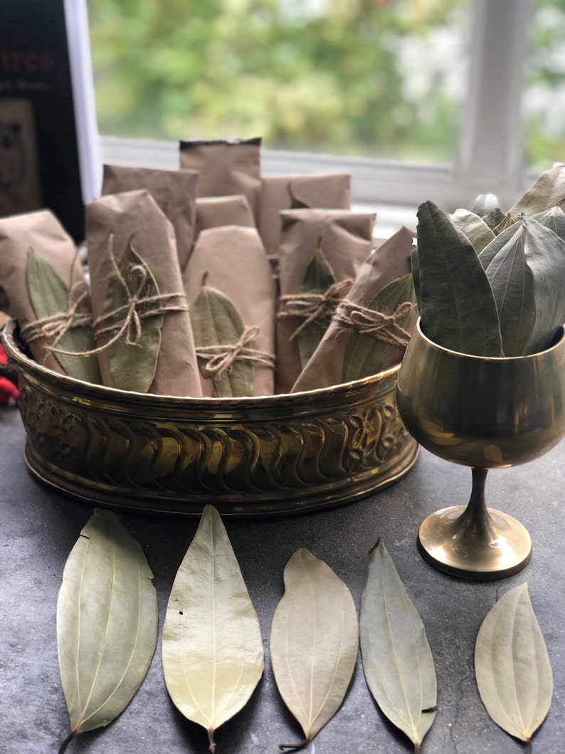 May include: A brass bowl filled with small brown paper packages tied with twine and decorated with dried bay leaves.  A gold goblet sits next to the bowl, also filled with dried bay leaves.  Several dried bay leaves are scattered on a gray surface.