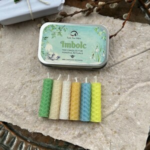 Może przedstawiać: Metalowa puszka z napisem "Imbolc Tiny Candles for Mindful Rituals" znajduje się nad sześcioma małymi, kolorowymi świecami z wosku pszczelego. Świece są zielone, kremowe, ż&oacute;łte, jasnoniebieskie i ż&oacute;łte. Puszka ma jasnoniebieski i zielony wz&oacute;r.