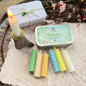 Może przedstawiać: Zapalone białe świeca w rustykalnym uchwycie, zapakowany prezent i puszka z napisem "Imbolc Tiny Candles for Mindful Rituals" są wyświetlane z kilkoma małymi, kolorowymi świecami. Świece są zielone, ż&oacute;łte, niebieskie i białe.