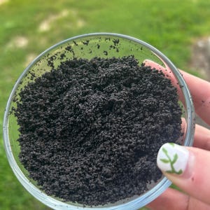 May include: A clear glass bowl filled with dark, granular material, possibly a spice or ground coffee. The bowl is held up against a blurred green background, suggesting an outdoor setting. The material appears to be finely ground and dark brown in color.
