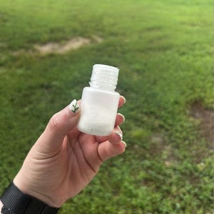 May include: A small, white, rectangular bottle with a white, ridged cap. The bottle appears to contain a white substance. The background is a blurred view of green grass.