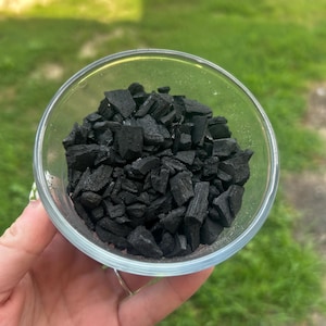 May include: A clear glass bowl filled with black charcoal pieces. The bowl is held in a hand, with a green grassy background. The charcoal pieces are irregularly shaped and have a matte finish.
