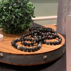 May include: A wooden display stand with a metal band and a sign that reads "Men's Bracelets $15.00".  Black and gray beaded bracelets are displayed on the stand.