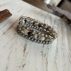 May include: A set of four bracelets stacked on top of each other. The bracelets are made of small, round beads in shades of brown, gray, and white. The beads are interspersed with clear, faceted beads. The bracelets are on a white, wooden surface.